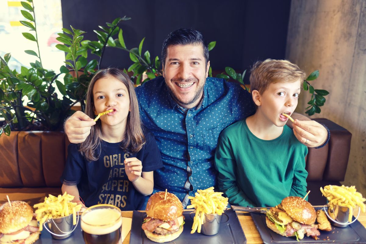 Which Holiday Foods are Braces-Safe Man and two children eating burgers and fries at table in casual indoor setting.